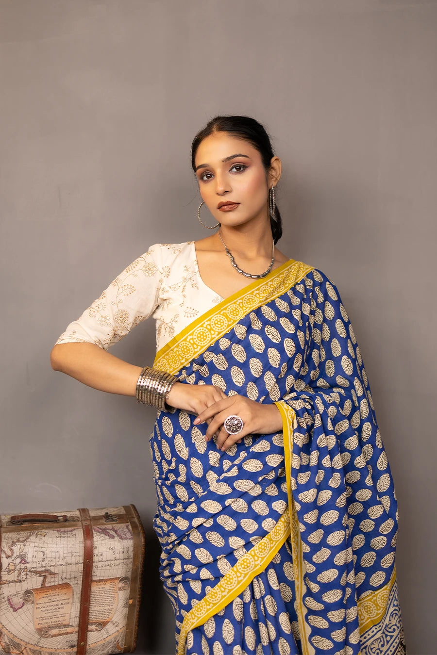 Woman wearing a blue and yellow saree standing next to vintage suitcases and books against a grey background