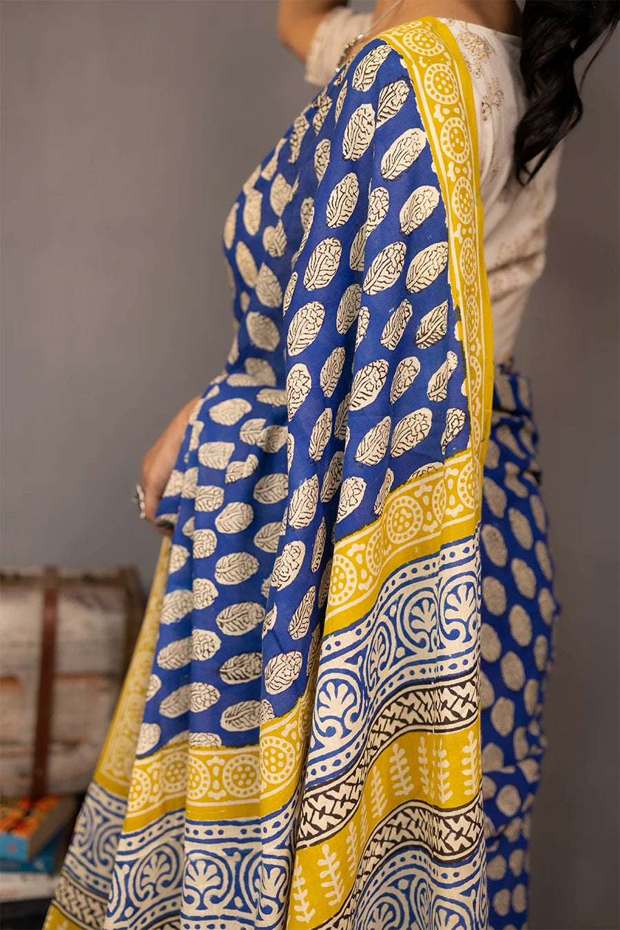 Woman wearing a blue and yellow saree standing next to vintage suitcases and books against a grey background