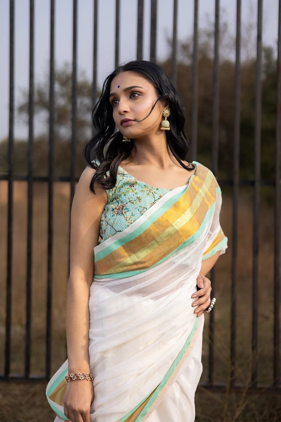Model wearing an off-white saree with gold and mint green striped border and tassel detailing, lightweight and suitable for festive and casual occasions.