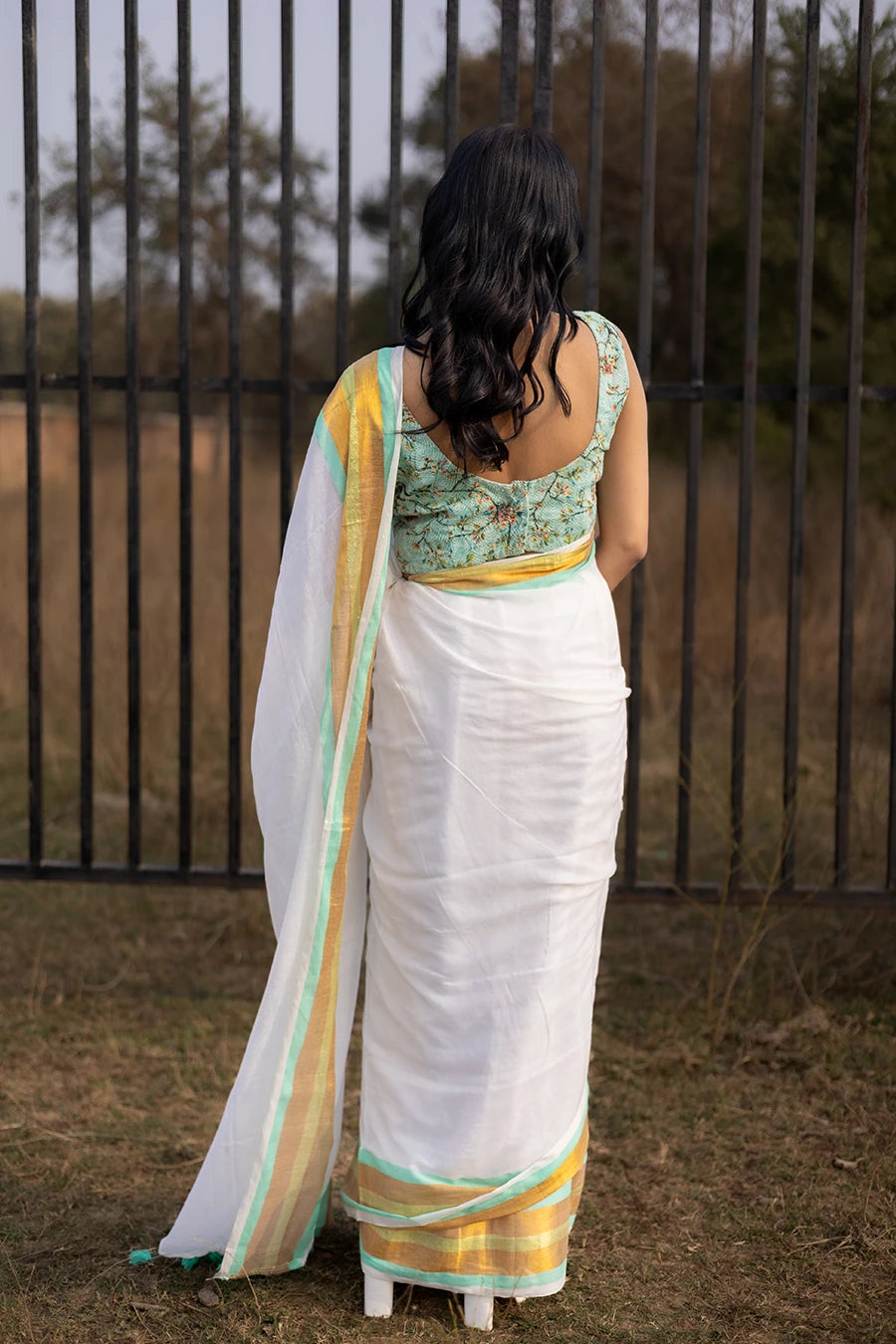 Model wearing an off-white saree with gold and mint green striped border and tassel detailing, lightweight and suitable for festive and casual occasions.