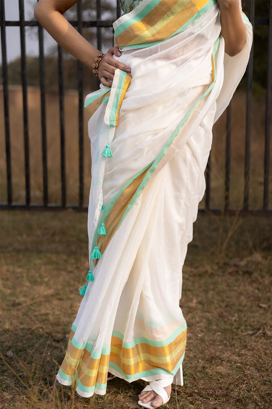 Model wearing an off-white saree with gold and mint green striped border and tassel detailing, lightweight and suitable for festive and casual occasions.