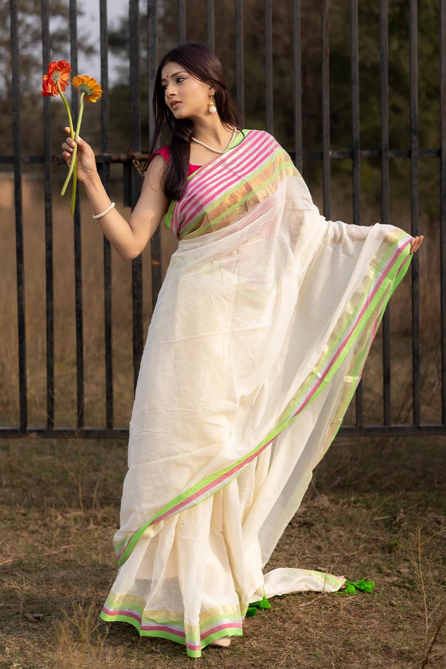 Model wearing an off-white saree with pink and green striped border and tassel detailing, lightweight and suitable for festive and casual occasions.