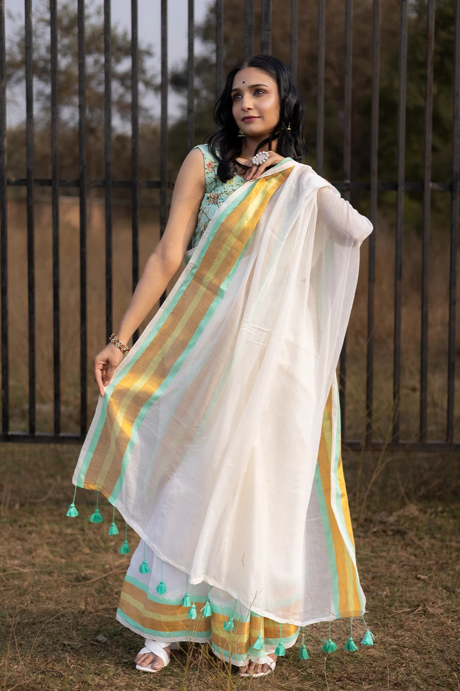 Model wearing an off-white saree with gold and mint green striped border and tassel detailing, lightweight and suitable for festive and casual occasions.