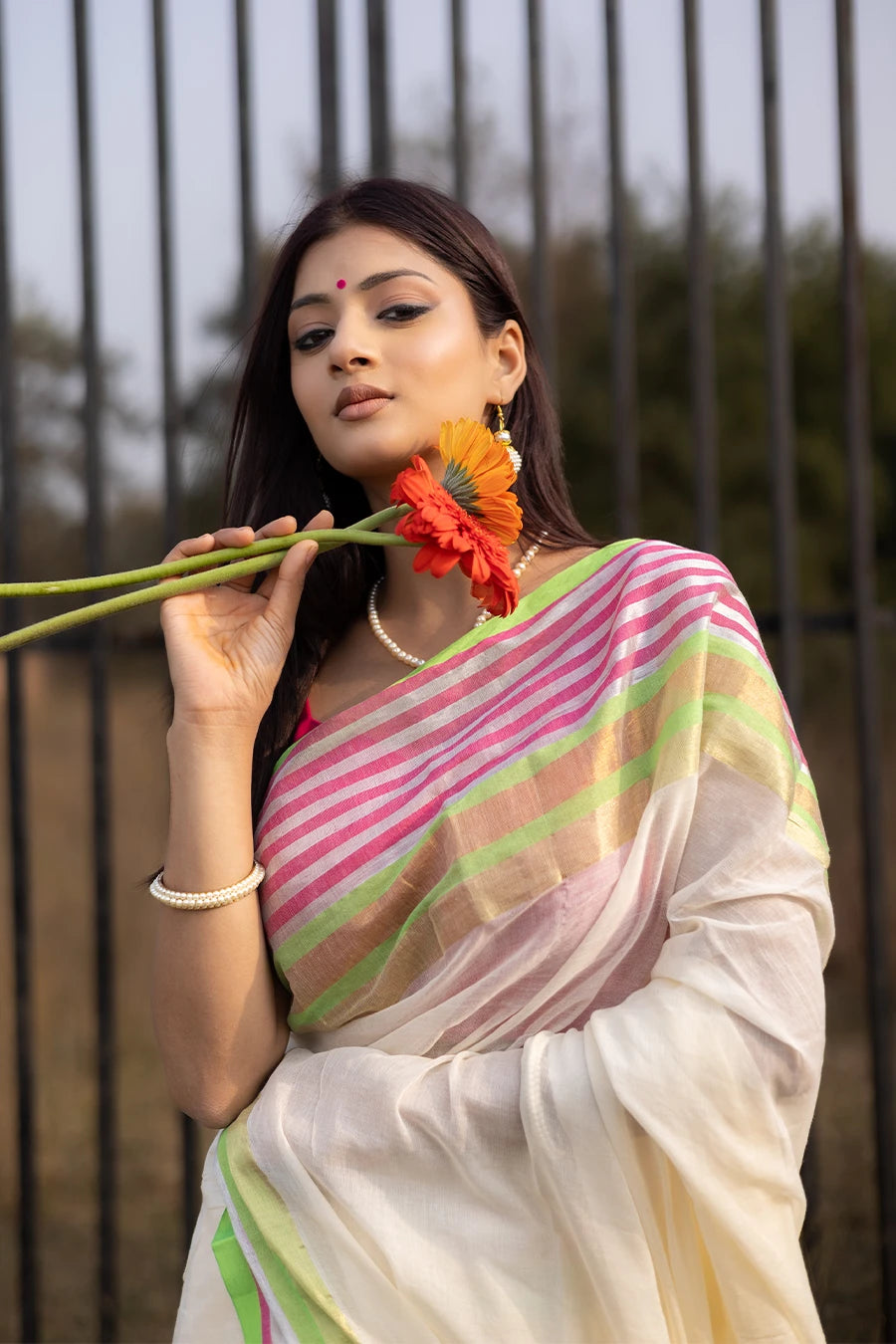 Model wearing an off-white saree with pink and green striped border and tassel detailing, lightweight and suitable for festive and casual occasions.