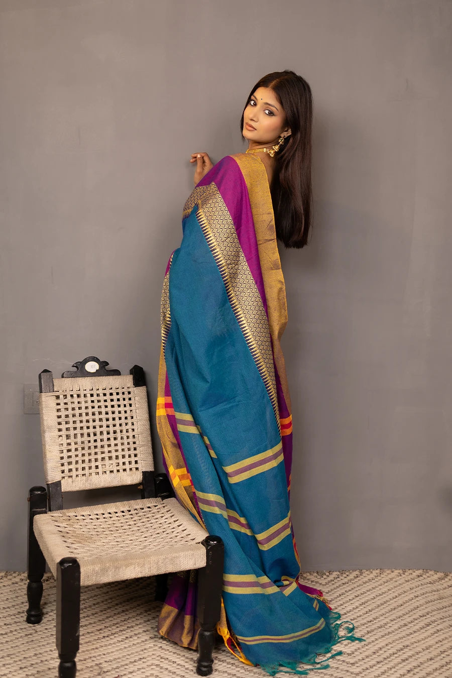 Woman wearing a blue saree with gold and purple accents, standing next to a chair against a grey background.