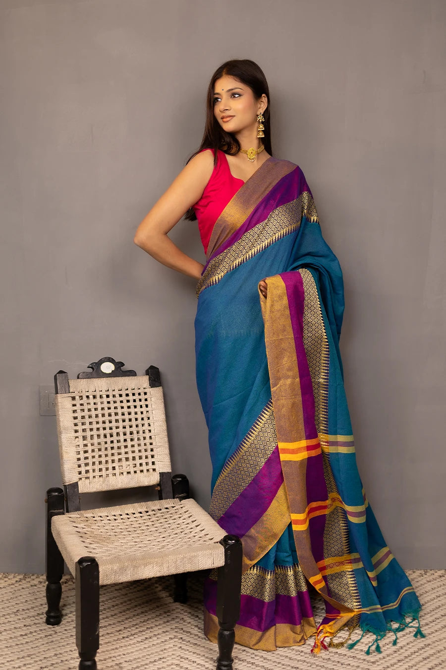 Woman wearing a blue saree with gold and purple accents, standing next to a chair against a grey background.