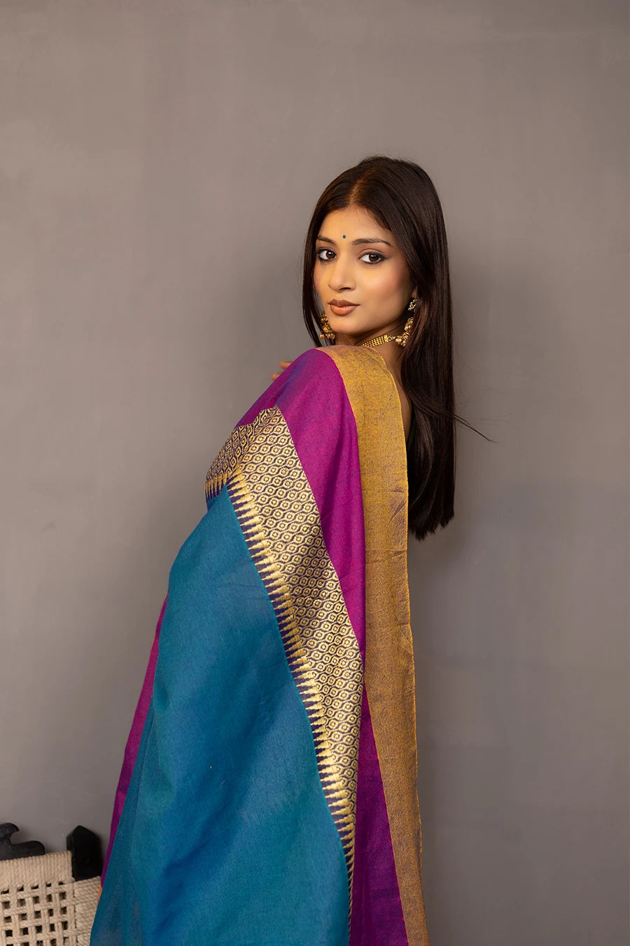 Woman wearing a blue saree with gold and purple accents, standing next to a chair against a grey background.