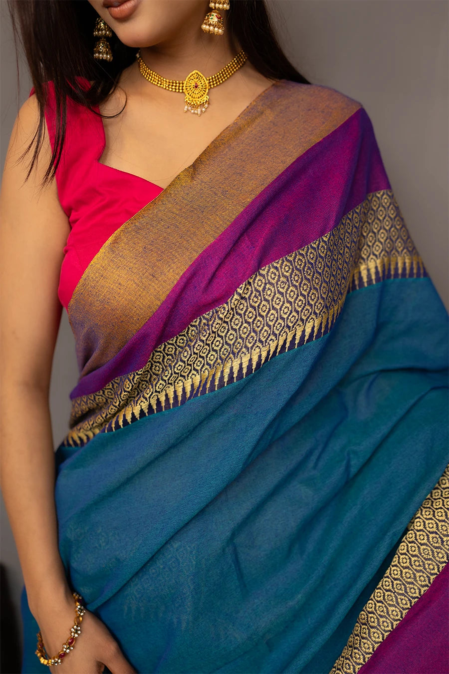 Woman wearing a blue saree with gold and purple accents, standing next to a chair against a grey background.