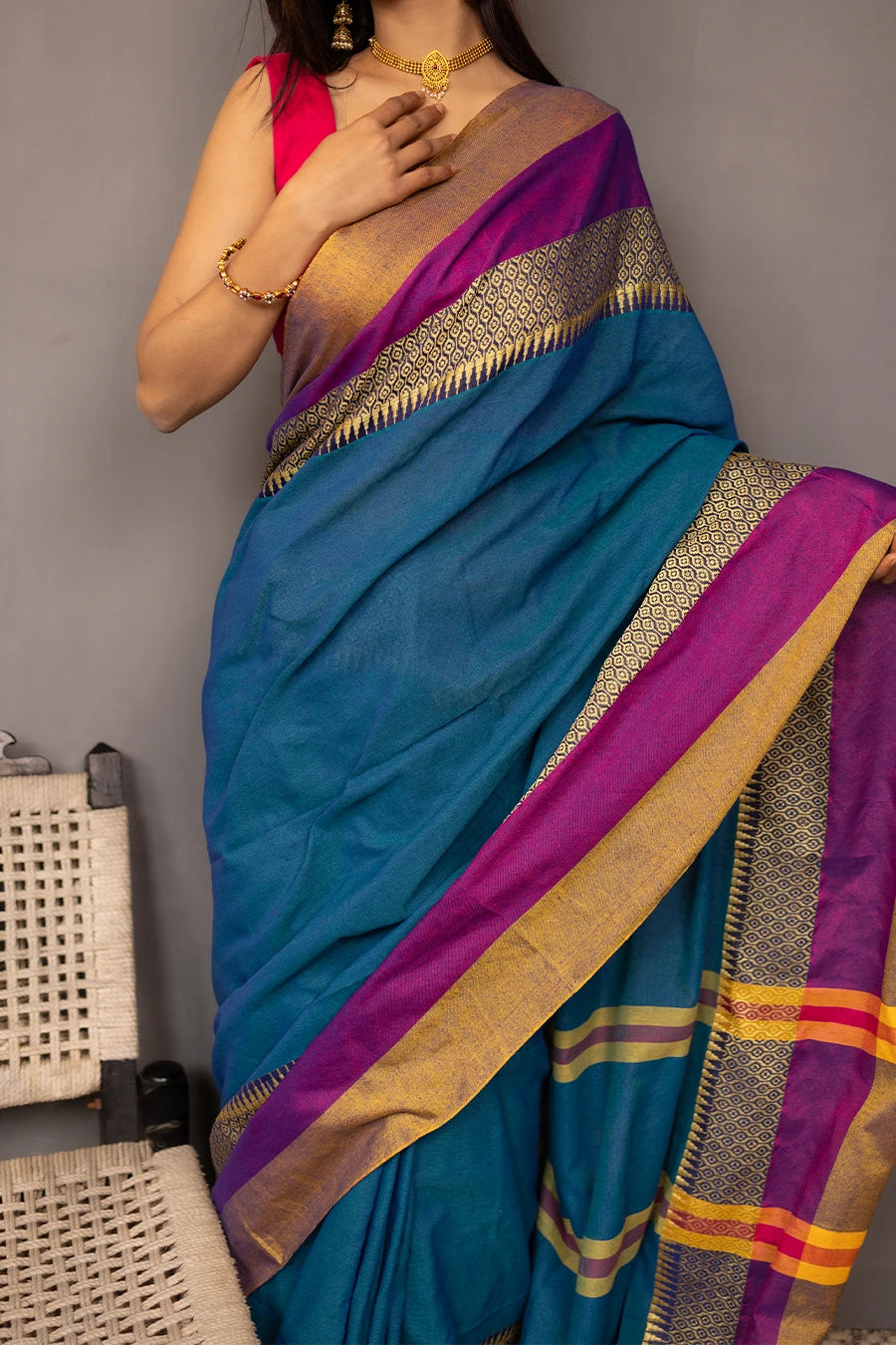 Woman wearing a blue saree with gold and purple accents, standing next to a chair against a grey background.