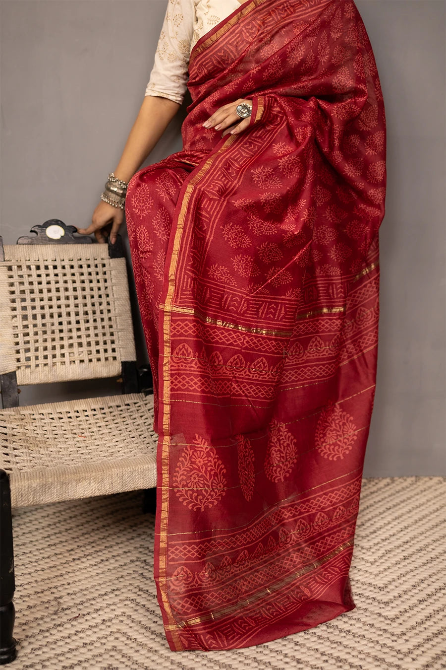 Woman in a red saree standing next to a chair against a grey wall.