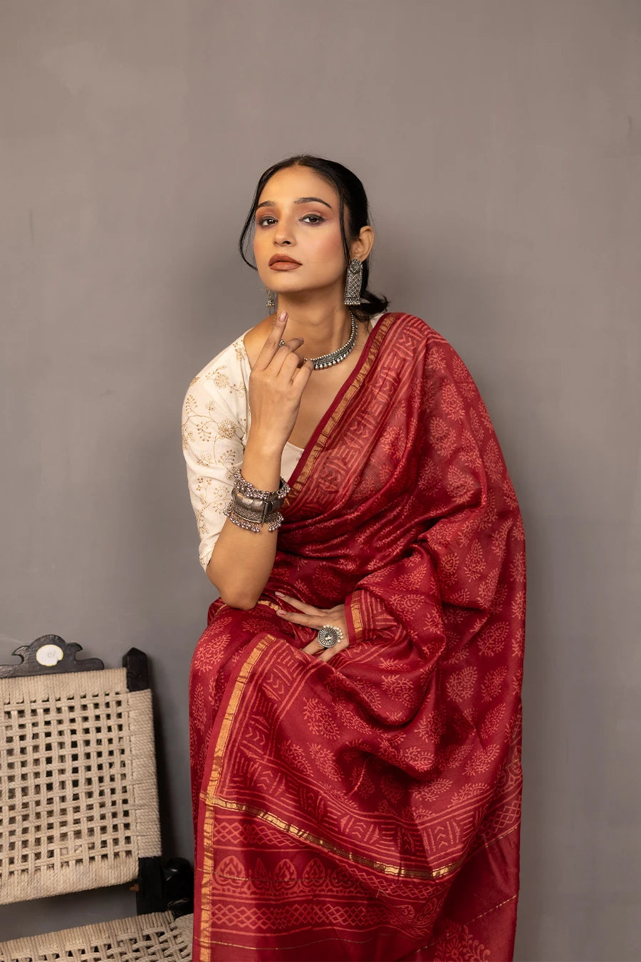 Woman in a red saree standing next to a chair against a grey wall.