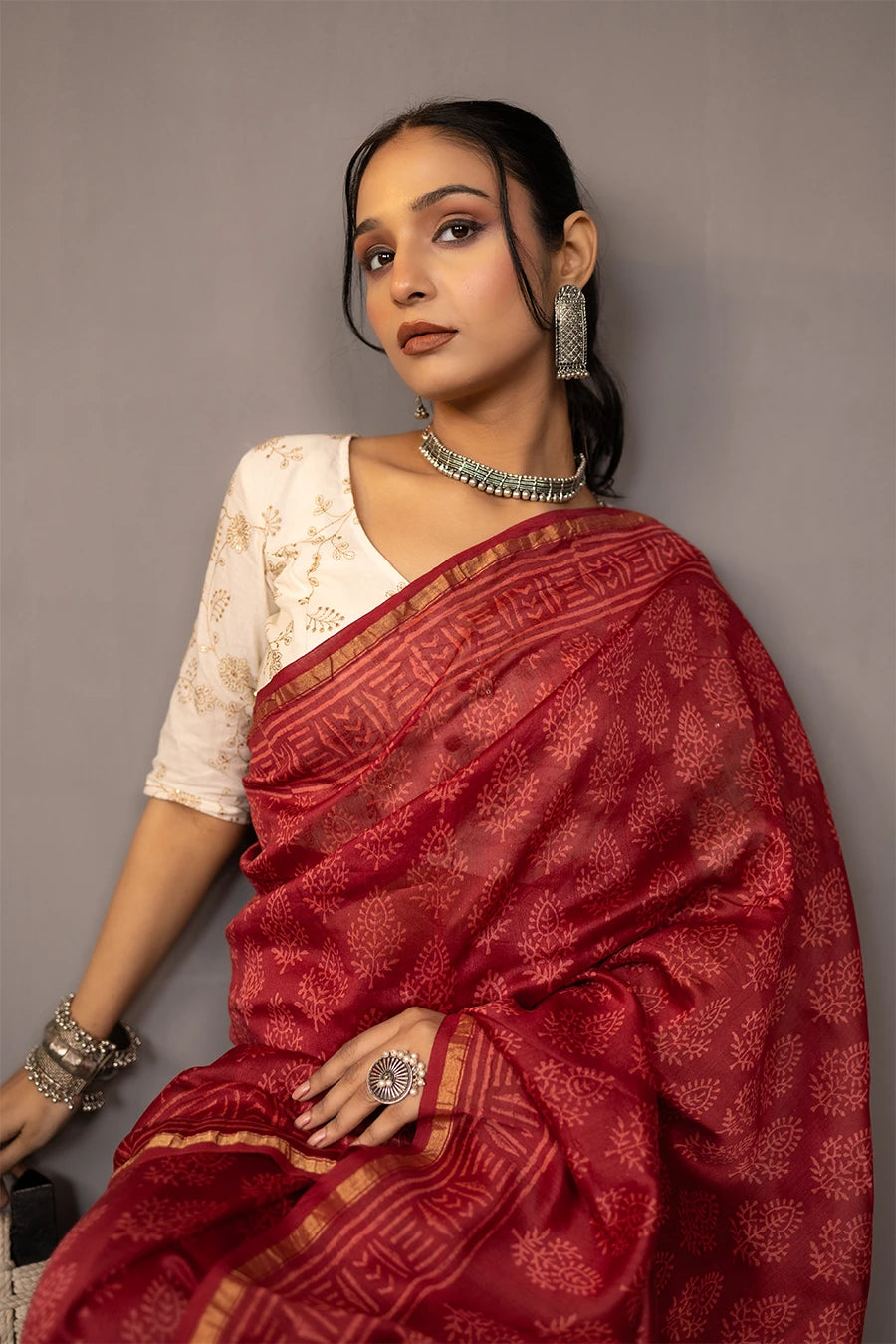 Woman in a red saree standing next to a chair against a grey wall.