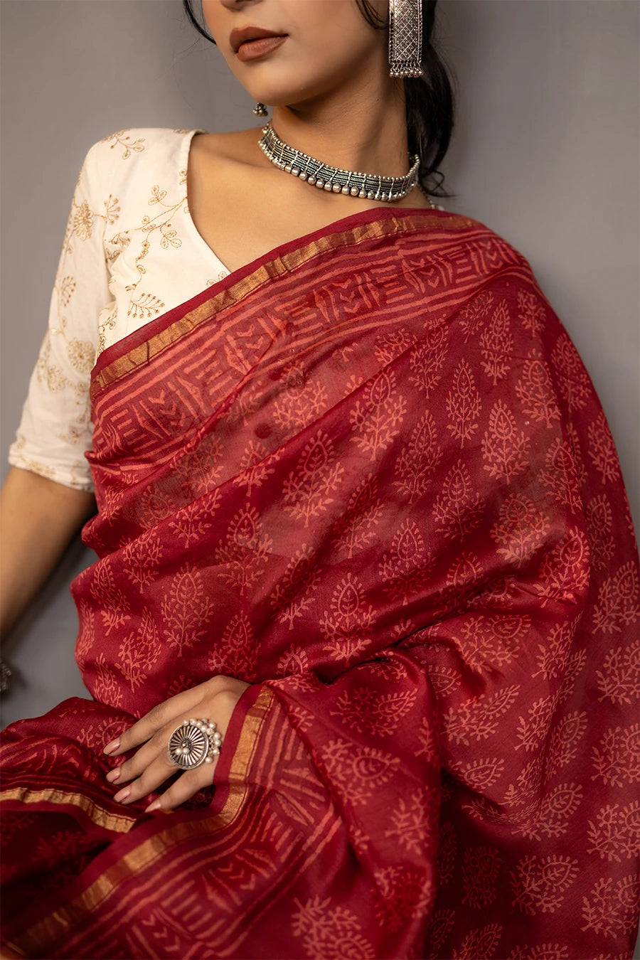 Woman in a red saree standing next to a chair against a grey wall.