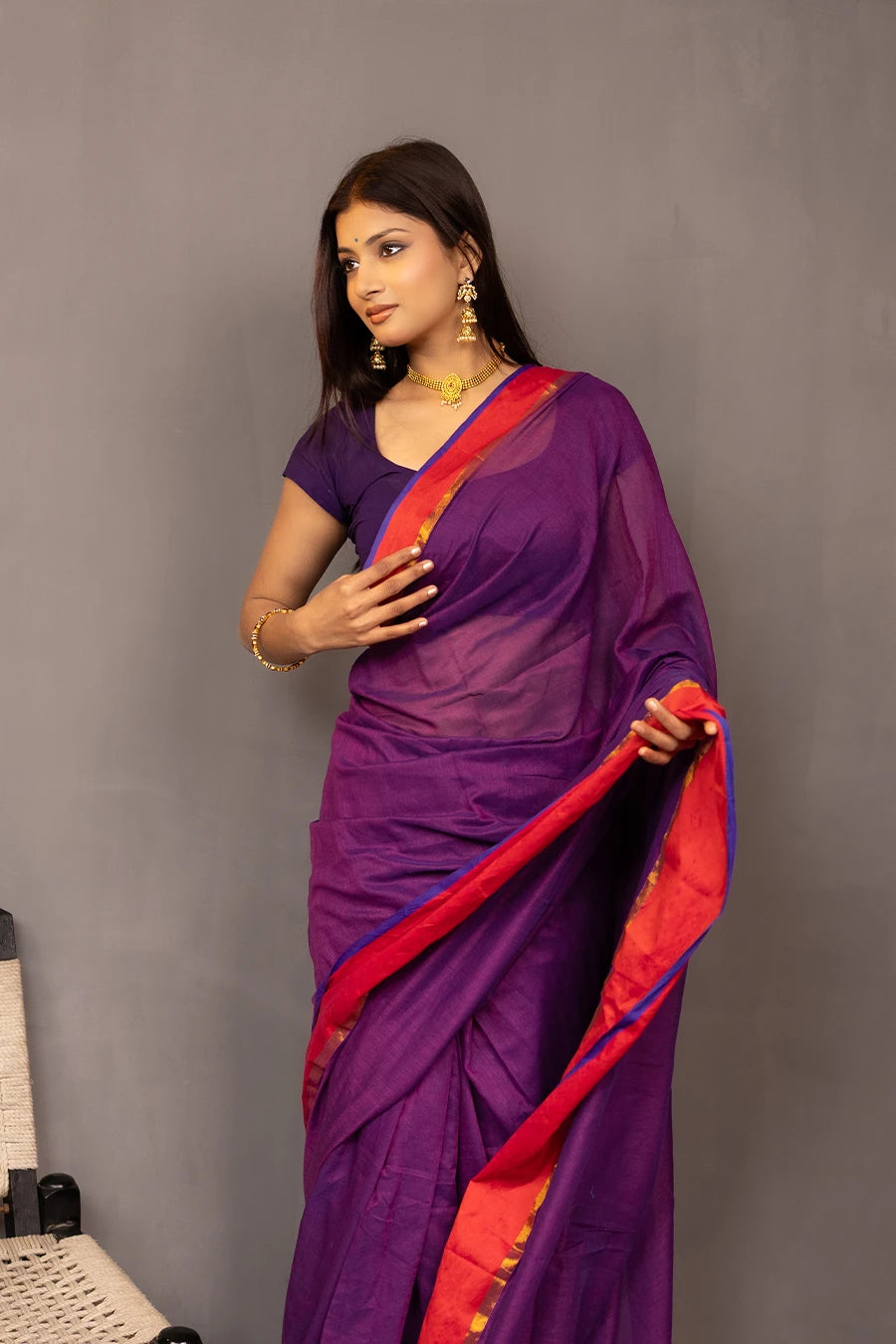 Woman wearing a purple saree with red border against a grey background