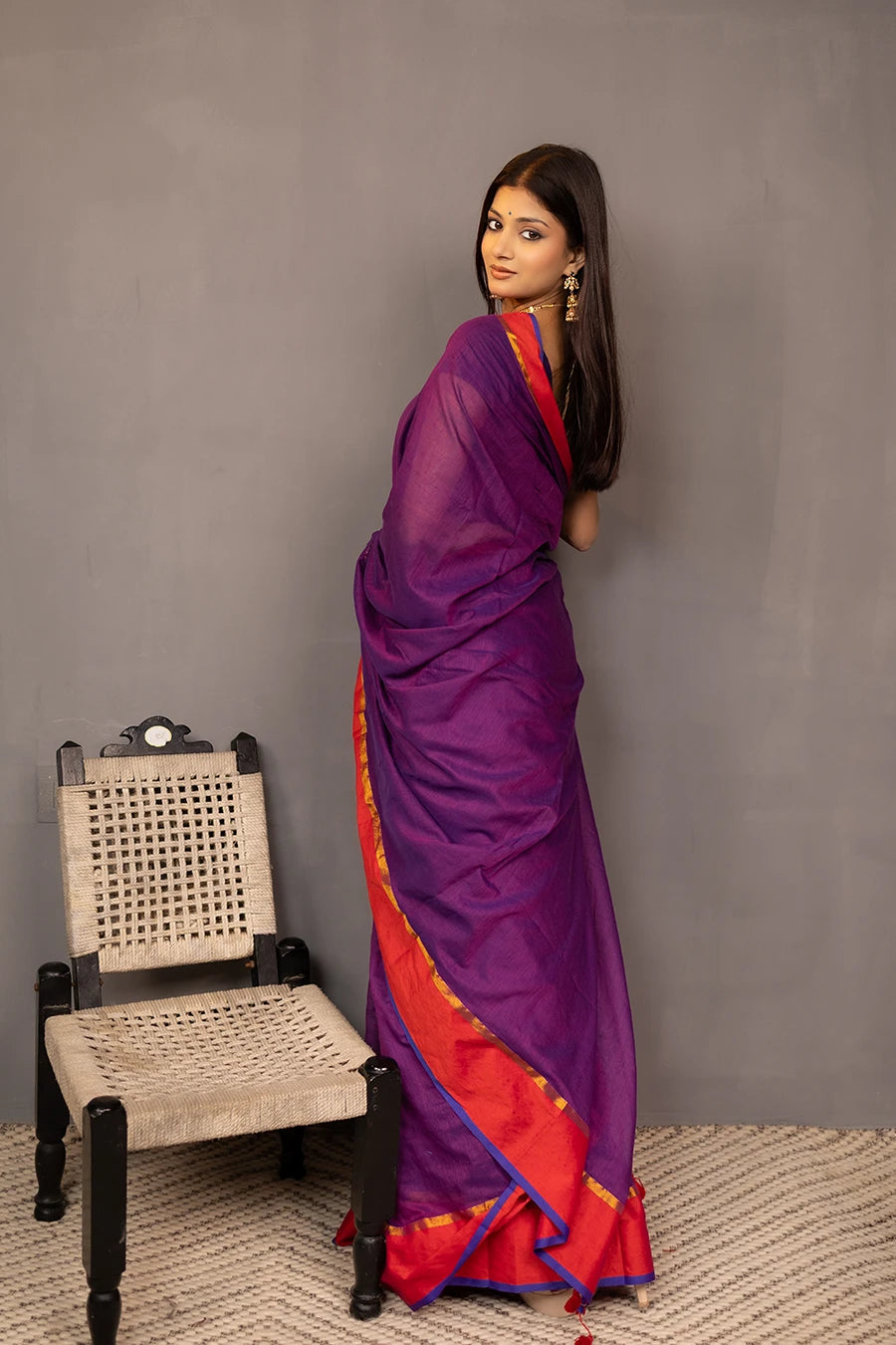 Woman wearing a purple saree with red border against a grey background