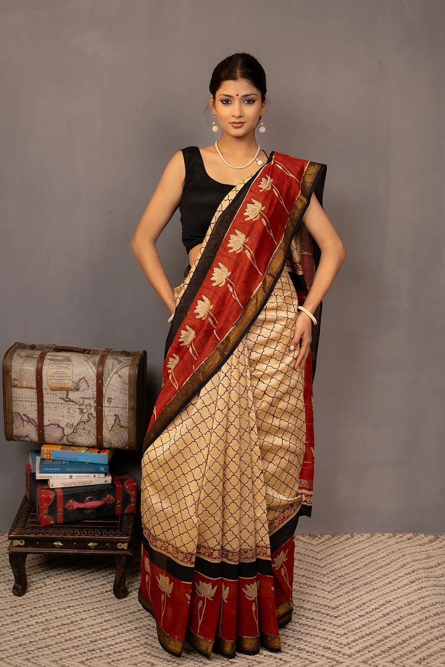 Woman wearing a traditional saree with a red border, standing next to a small table with books and a map.