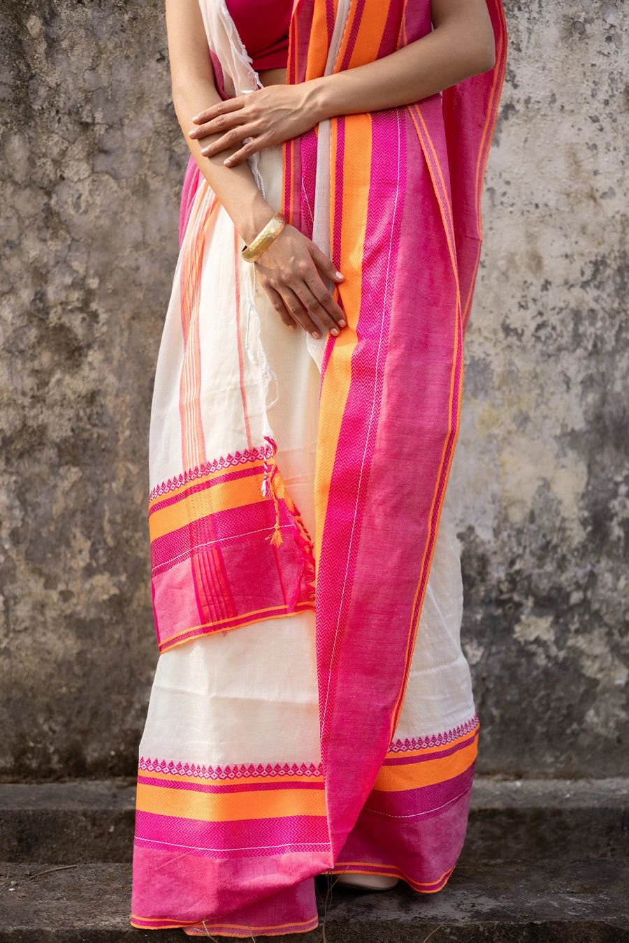 Woman wearing a traditional saree with pink and orange border against a textured wall.