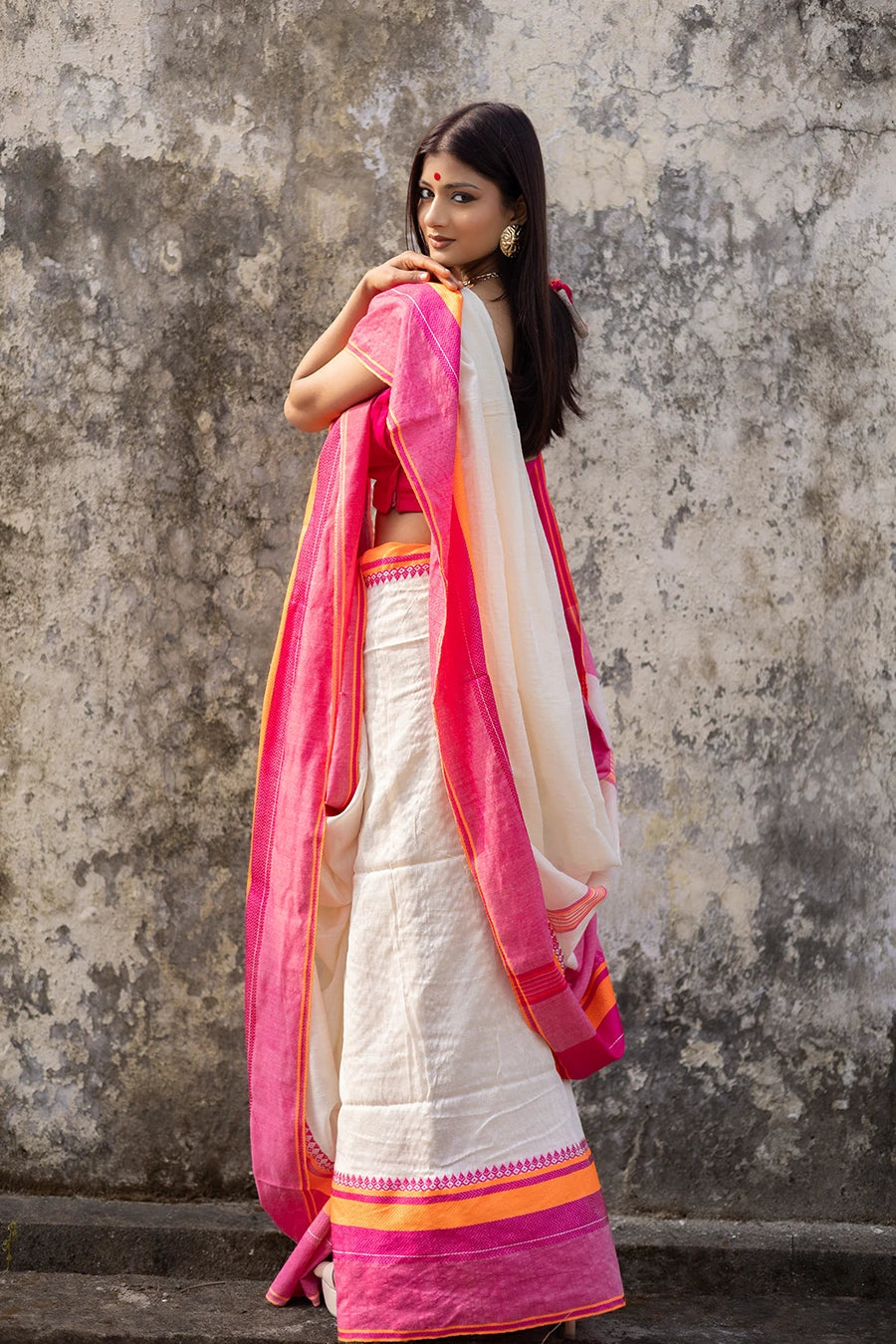 Woman wearing a traditional saree with pink and orange border against a textured wall.