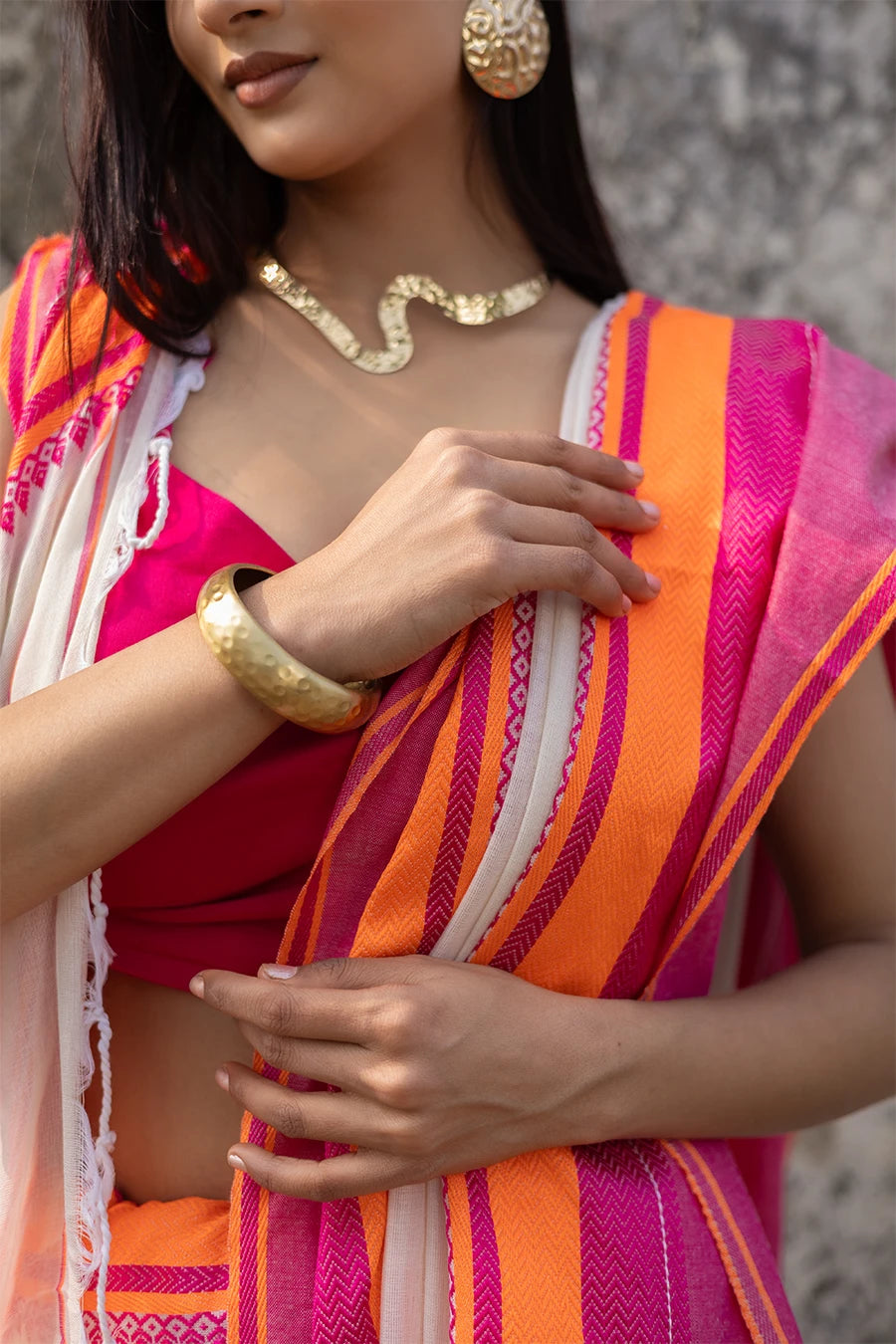 Woman wearing a traditional saree with pink and orange border against a textured wall.