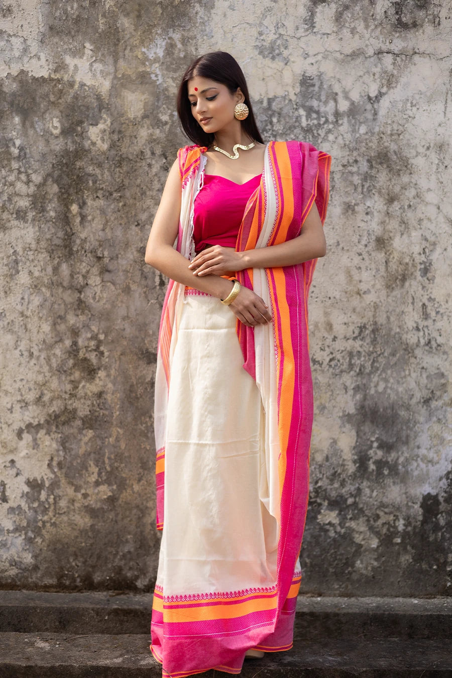 Woman wearing a traditional saree with pink and orange border against a textured wall.