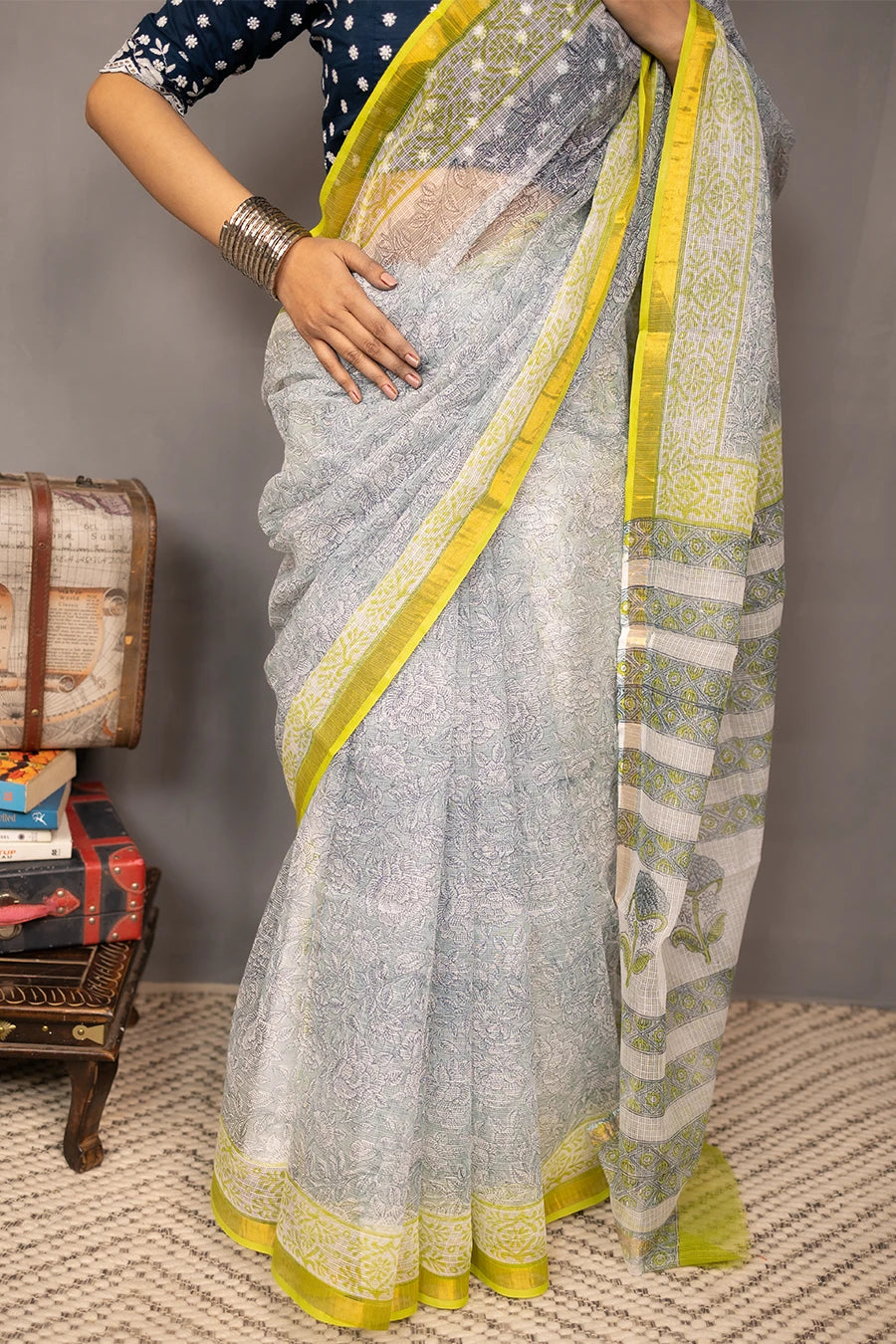 Woman in a saree standing next to a small table with books and boxes against a grey background