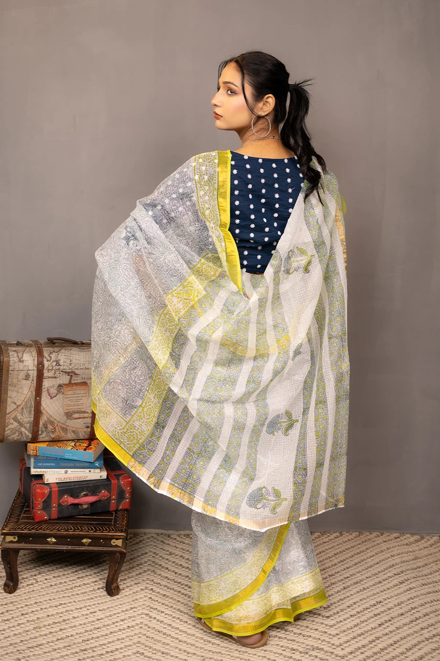 Woman in a saree standing next to a small table with books and boxes against a grey background