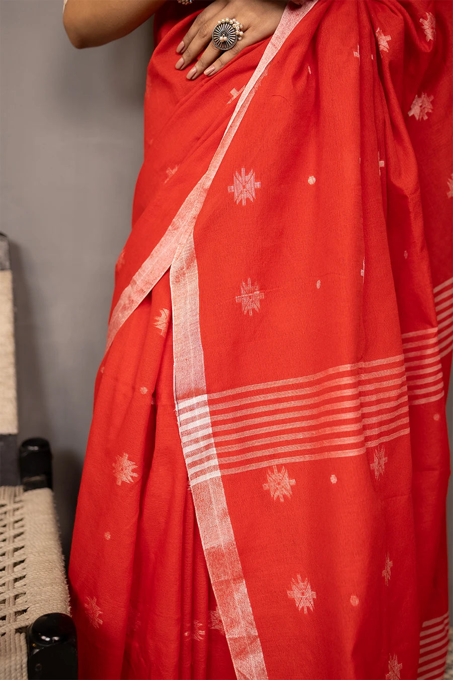 Woman in a red saree standing next to a chair against a grey wall