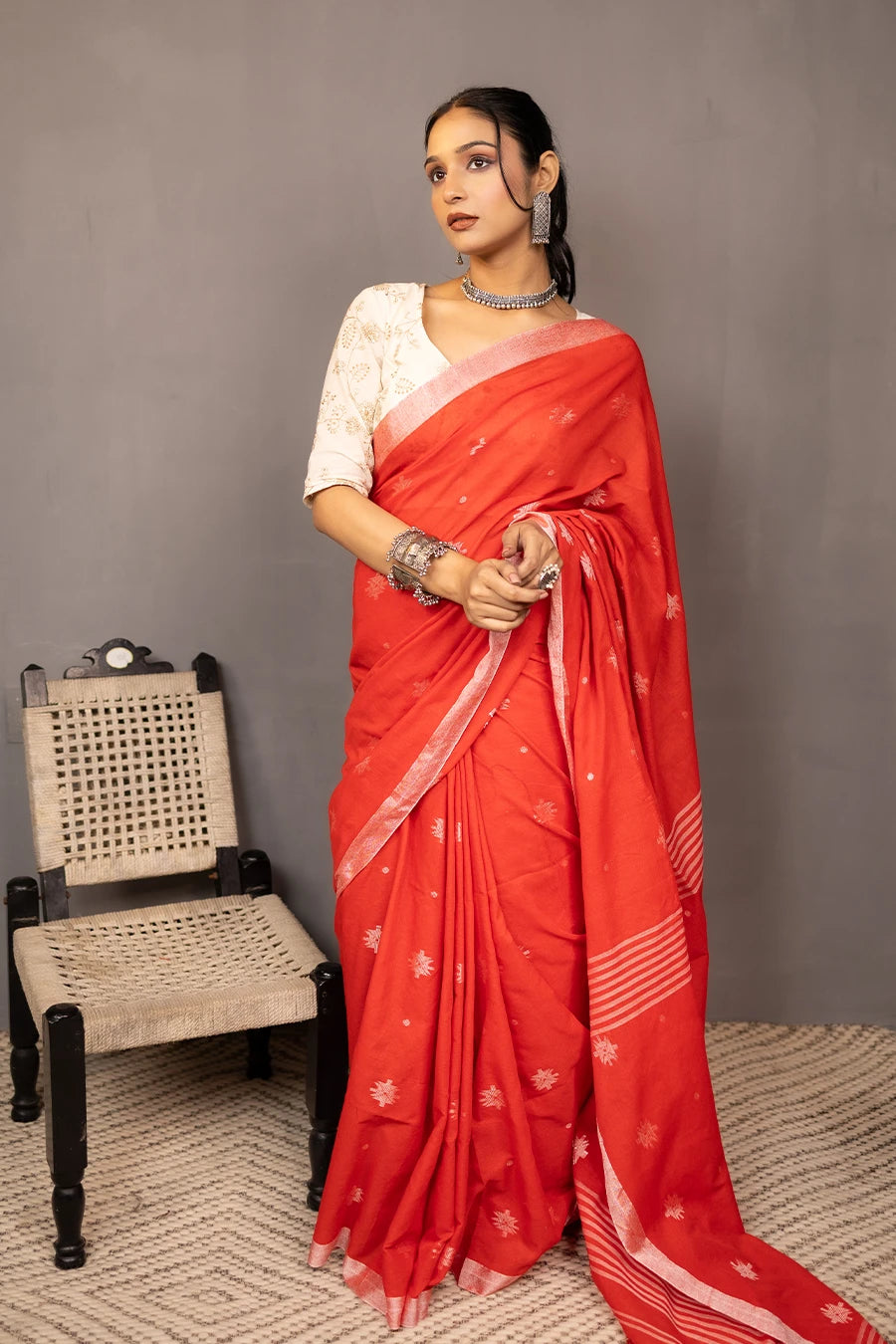 Woman in a red saree standing next to a chair against a grey wall