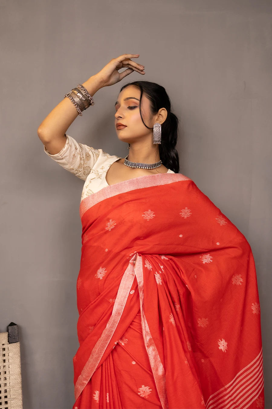 Woman in a red saree standing next to a chair against a grey wall
