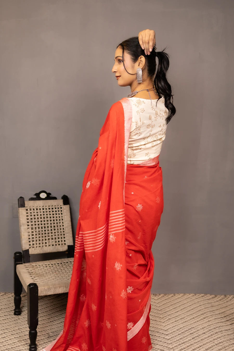 Woman in a red saree standing next to a chair against a grey wall