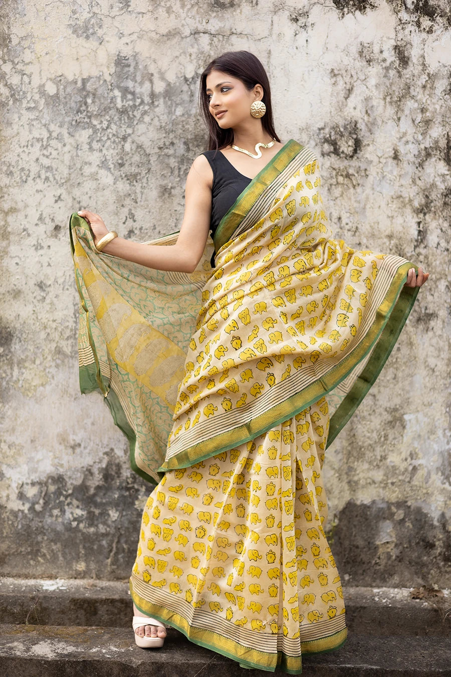 Woman in a yellow and green saree standing against a textured wall.