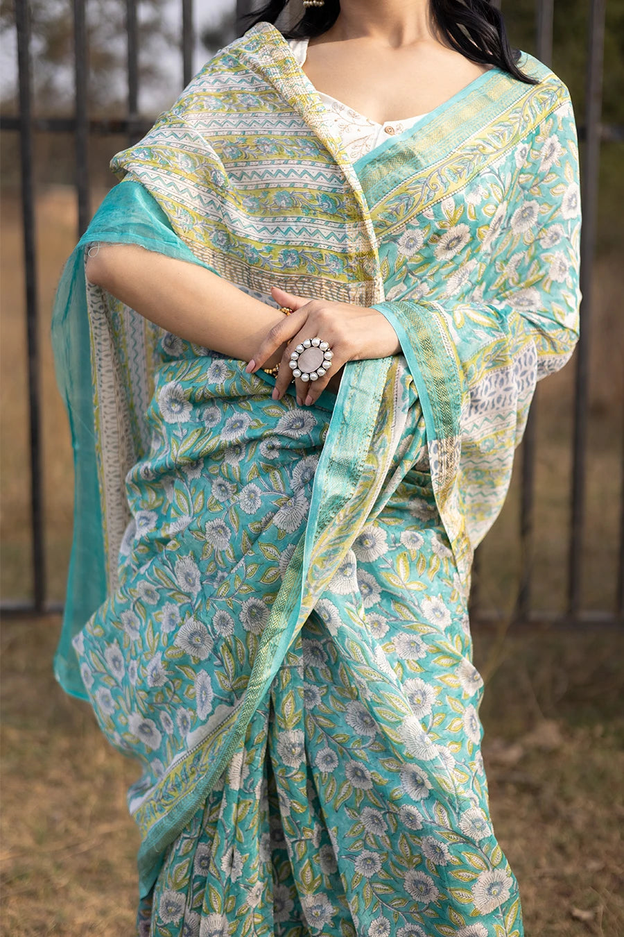 Woman wearing a green floral saree standing outdoors with a metal fence and grass in the background.