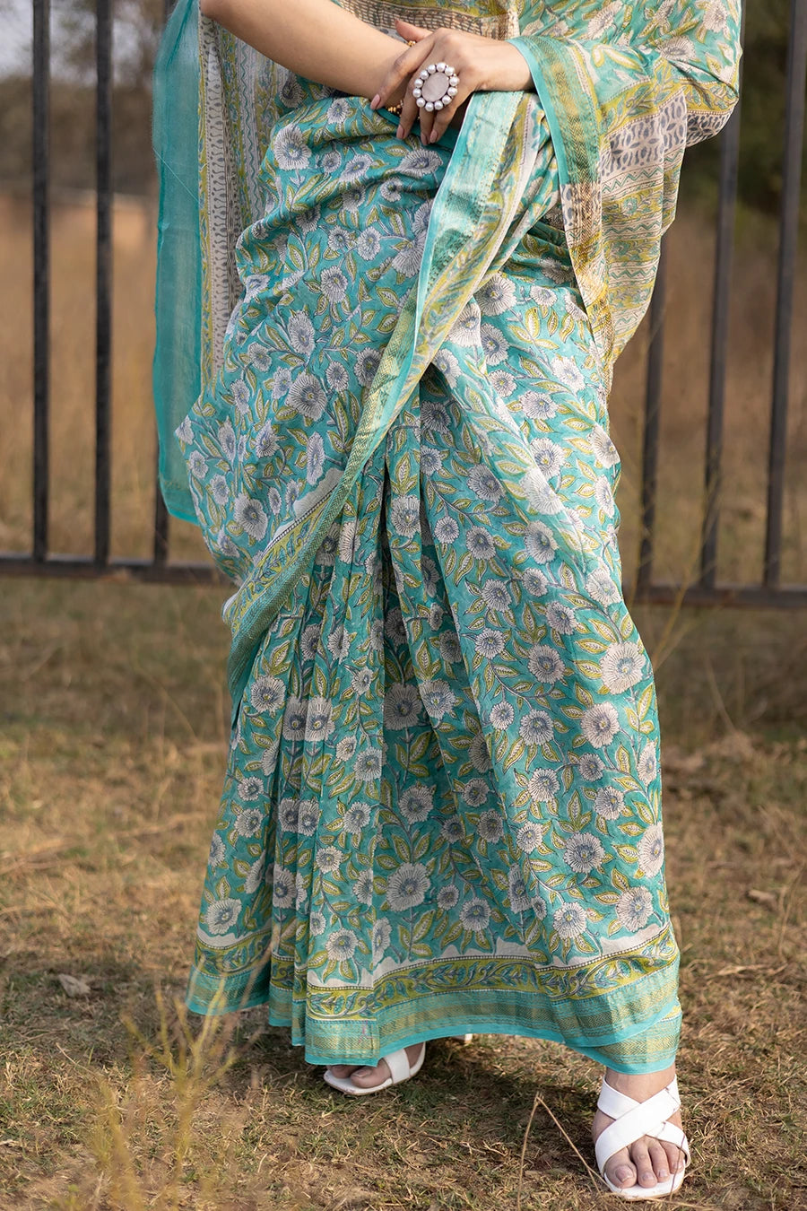 Woman wearing a green floral saree standing outdoors with a metal fence and grass in the background.