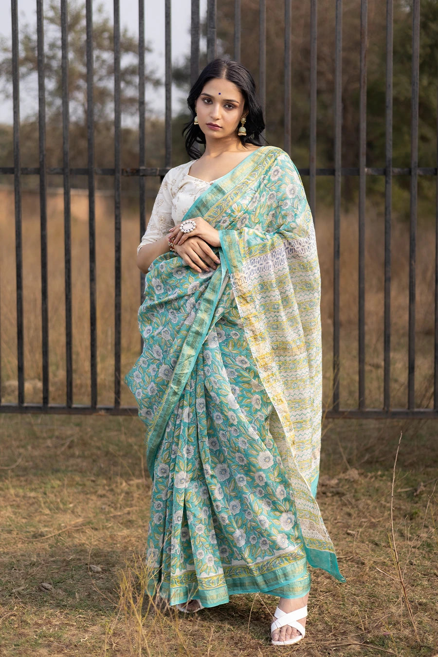 Woman wearing a green floral saree standing outdoors with a metal fence and grass in the background.