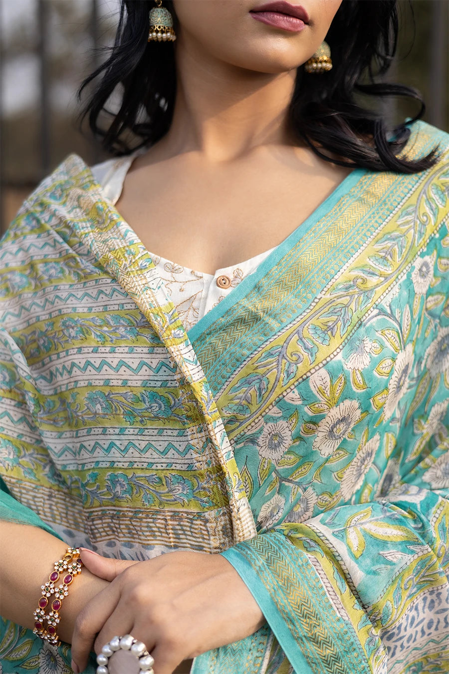 Woman wearing a green floral saree standing outdoors with a metal fence and grass in the background.