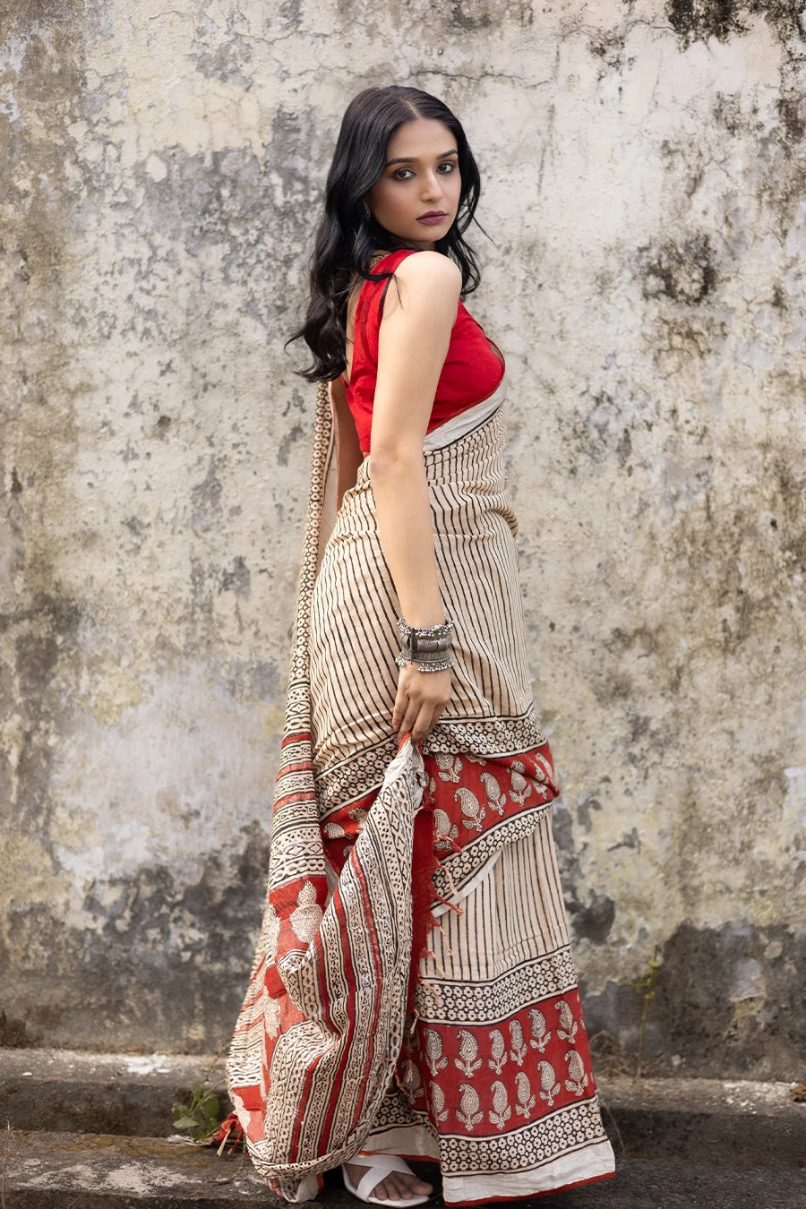Woman in a red and beige saree standing on steps with a textured wall background