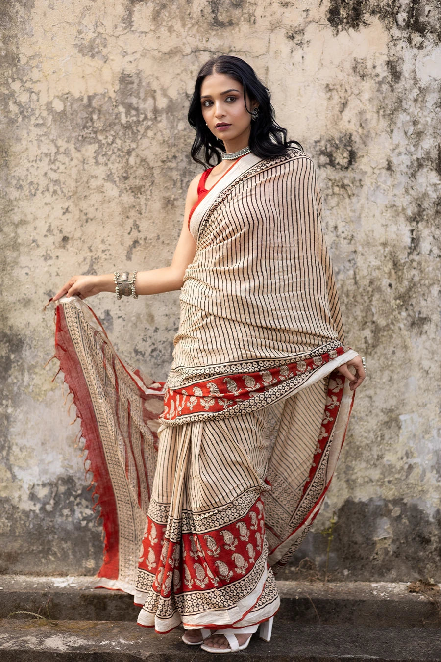 Woman in a red and beige saree standing on steps with a textured wall background