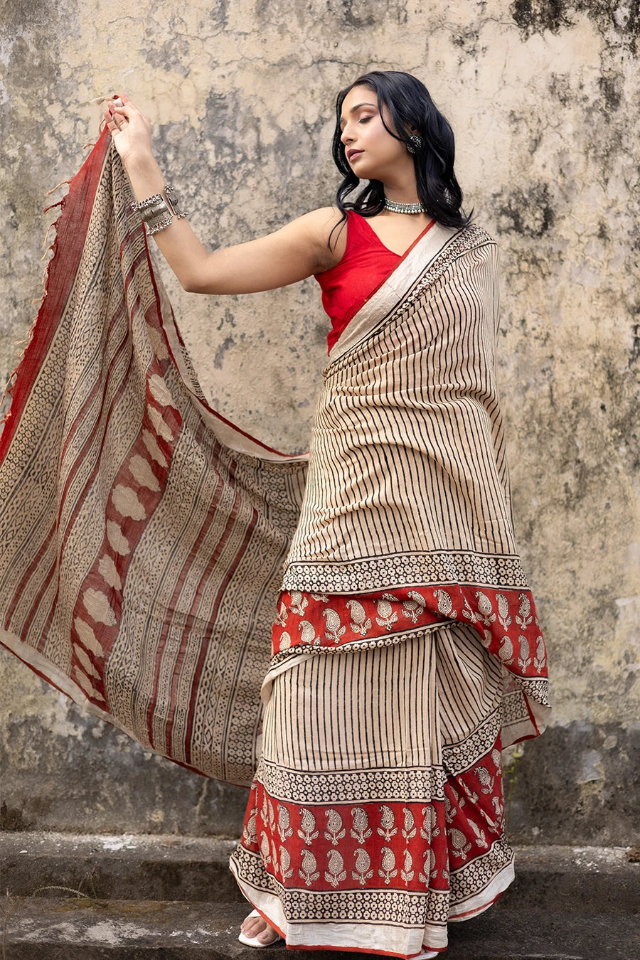 Woman in a red and beige saree standing on steps with a textured wall background