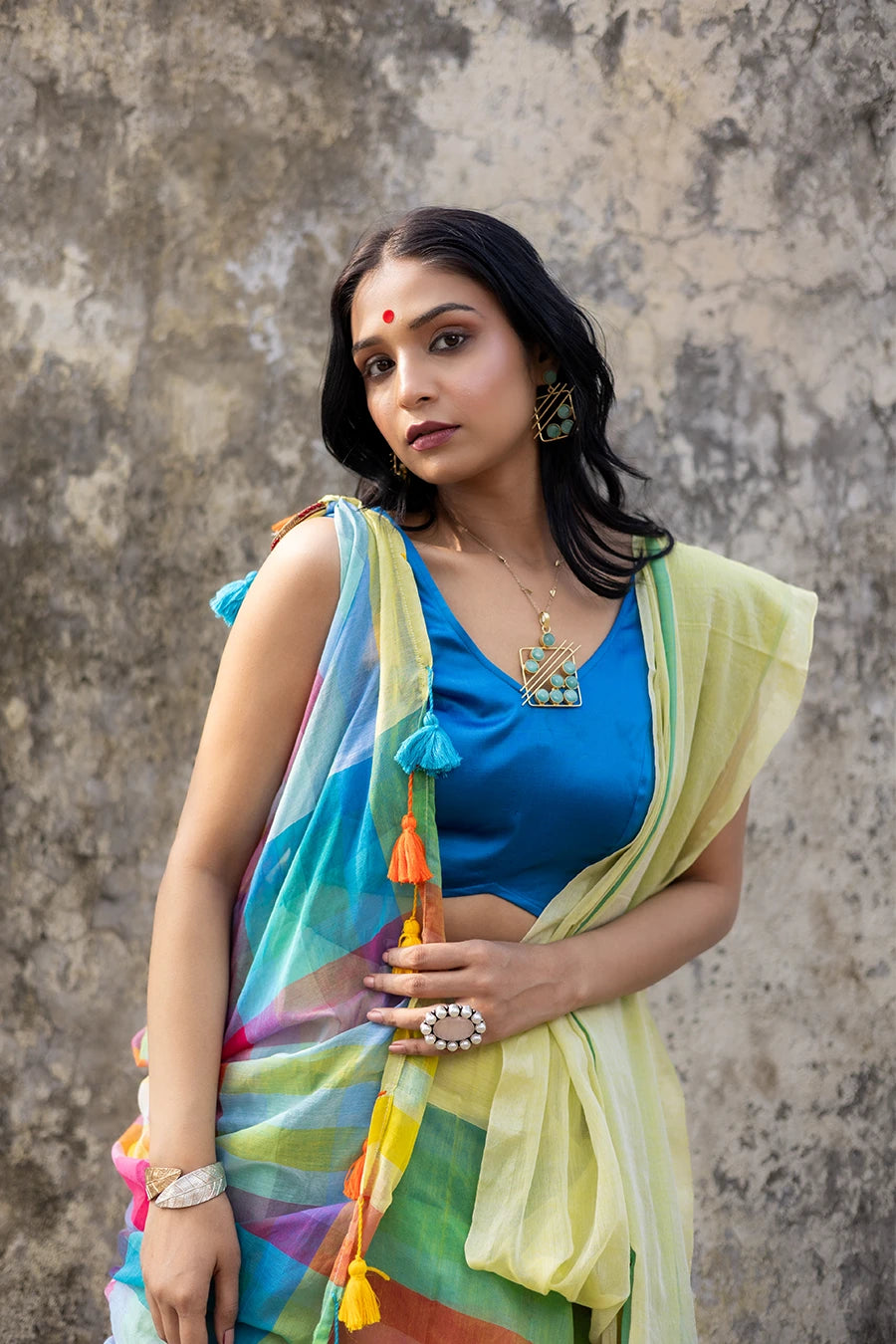 Woman wearing a colorful saree with a blue blouse against a textured wall.